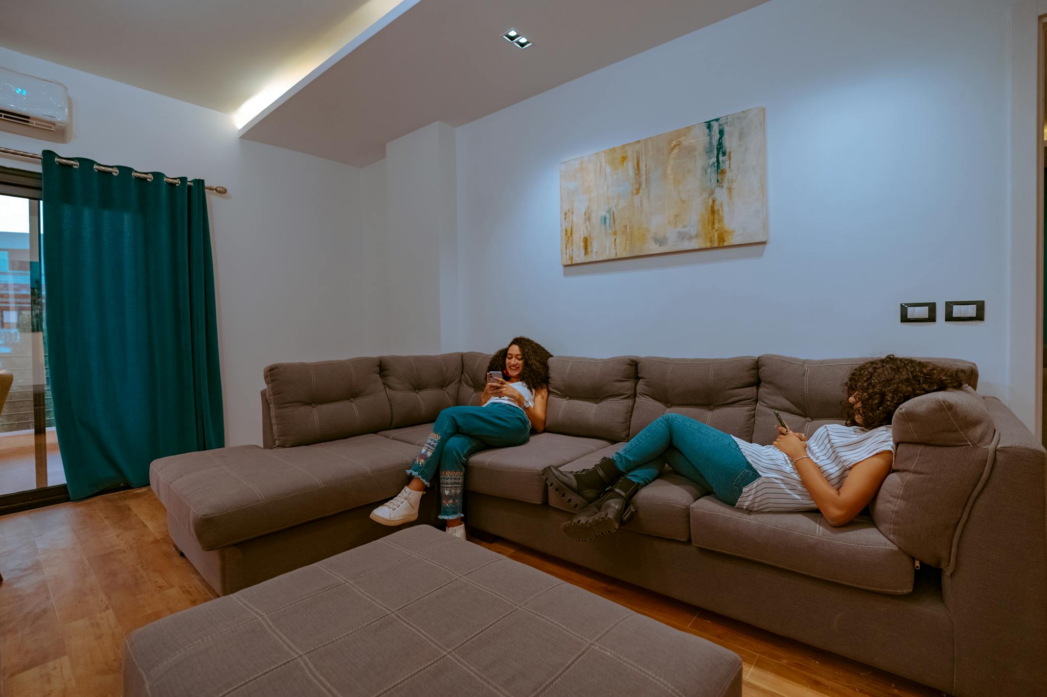 Two women with curly hair relaxing on a sofa, using smartphones in a modern living room.